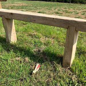 May include: A rustic wooden bench with two legs on each side, made from rough-hewn timber. The bench is set in a grassy field.
