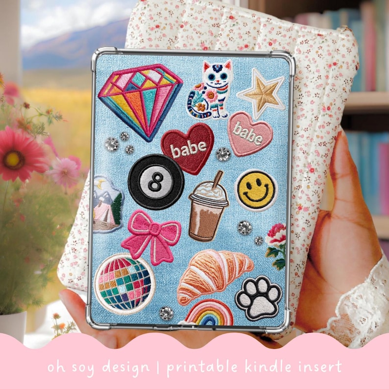 May include: A light blue Kindle case with various embroidered patches, including a diamond, cat, star, hearts with "babe" text, an 8-ball, coffee drink, smiley face, bow, disco ball, croissant, rainbow, and paw print. The text "oh soy design | printable kindle insert" is at the bottom.