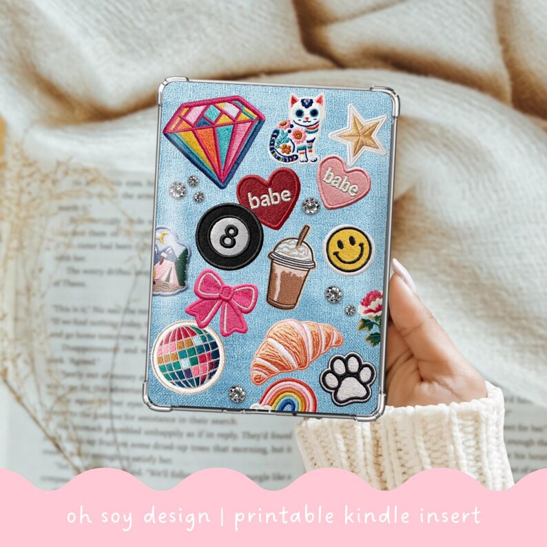 May include: A light blue Kindle insert with various embroidered patches, including a rainbow diamond, a cat, hearts, an eight ball, a bow, a disco ball, a croissant, a rainbow, a paw print, and a smiley face. The text "oh soy design | printable kindle insert" is at the bottom.