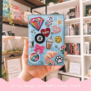 May include: A light blue Kindle insert decorated with colorful embroidered patches, including a diamond, hearts with "babe" text, a cat, a star, a smiley face, and a croissant. The insert also features a bow, a rainbow, and a paw print. The text "oh soy design | printable kindle insert" is at the bottom.