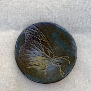 Puede incluir: Un plato redondo de cerámica con un esmalte verde oscuro y un diseño de mariposa en un esmalte marrón más claro.