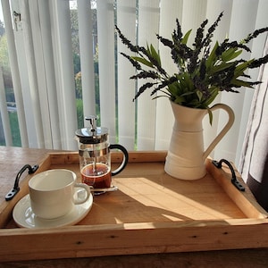 May include: A rectangular wooden serving tray with black metal handles containing a glass French press with coffee, a white ceramic teacup on a matching saucer, and a white ceramic pitcher filled with dark purple artificial flowers.