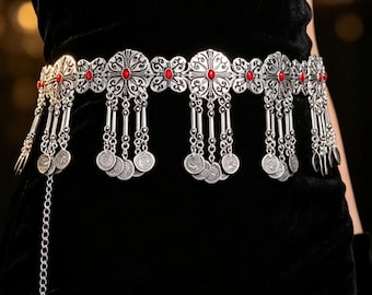 Pode incluir: Um cinto de cintura em metal prateado com uma série de elos florais ornamentados, cada um centrado com uma pequena pedra oval vermelha. O cinto é adornado com correntes prateadas pendentes e berloques circulares tipo moeda.