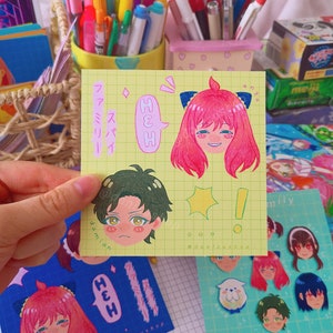May include: A yellow sheet of paper with stickers on it. The stickers feature cartoon characters with speech bubbles that say "HEH" and "anya". The characters are drawn in a colorful anime style. The sheet of paper has a grid pattern.