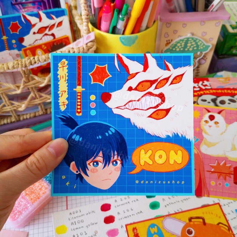 May include: A blue and white illustrated card with a white wolf with red eyes and a blue-haired boy with a red sword. The card has the text "KON" in yellow and "@danizushop" in black.