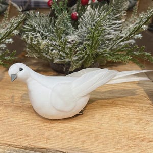 May include: A white dove ornament with detailed feathering and a slightly open beak. The bird sits on a wooden surface, with frosted greenery and red berries in the background, suggesting a festive theme.