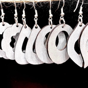 Op de afbeelding: Een set van acht zilver geverfde houten oorbellen met verschillende uitgesneden vormen. De oorbellen hangen aan zilveren haakjes.