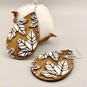 Puede incluir: Pendientes de madera en forma de lágrima con diseño de hojas. Los pendientes son marrones con recortes de hojas blancas y ganchos plateados. Se muestran sobre un fondo blanco.