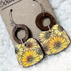 May include: A pair of wooden earrings with a sunflower design. The earrings are made of two pieces of wood, one round and one square. The square piece is decorated with a pattern of sunflowers in shades of yellow and orange. The earrings are attached to silver hoops.