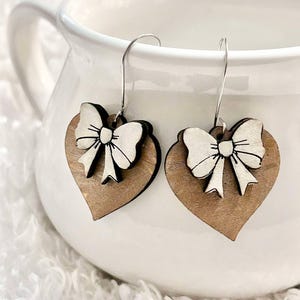 May include: A pair of wooden heart-shaped earrings with white bow accents. The earrings are hanging from a white ceramic mug.