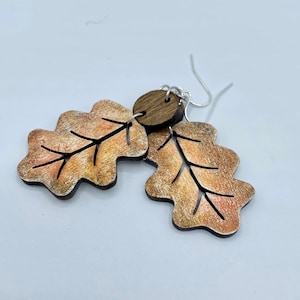 Puede incluir: Un par de pendientes de madera con forma de hojas de roble con un acento de madera marrón. Las hojas están pintadas con un degradado de naranja, amarillo y marrón.