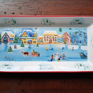 Puede incluir: Una bandeja rectangular de servir de cerámica blanca con un borde rojo. La bandeja presenta una escena de pueblo de invierno con casas coloridas, árboles y personas. La escena está pintada en un estilo caprichoso.