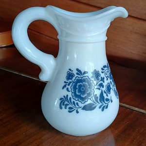 May include: White milk glass pitcher with a curved handle and spout. The pitcher features a blue floral design on the front. The pitcher is a vintage style and is likely used for serving beverages.