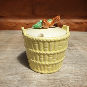 May include: A yellow ceramic sugar bowl with a lid and a handle. The bowl is designed to look like a woven basket. The lid has a small handle with a brown branch and green leaves. The bowl is sitting on a wooden surface.