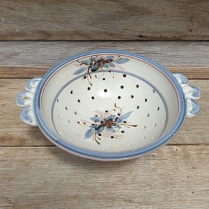 May include: A white ceramic colander with blue and pink floral designs and two loop handles. The colander has a white interior with small holes for draining.