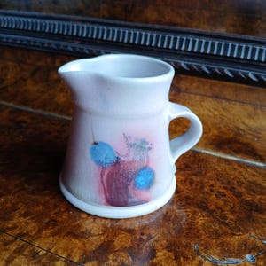 Pode incluir: Um jarro de cerâmica branca com uma alça. O jarro tem um design abstrato rosa e azul.