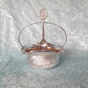 May include: Silver sugar bowl with a lid and a matching spoon. The bowl has a decorative handle and the spoon has a floral design.