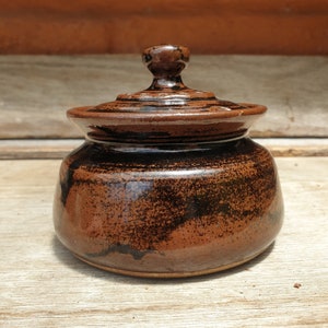 May include: A small, brown ceramic jar with a lid. The jar has a speckled brown glaze with black accents. The lid has a rounded knob.
