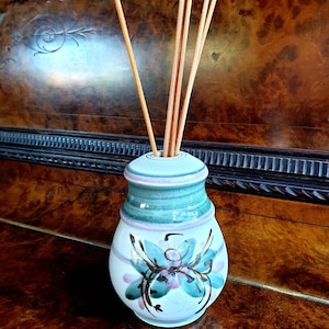 Może przedstawiać: Biała ceramiczna butelka dyfuzora z zielonym i różowym wzorem kwiatowym. Butelka ma okrągłą podstawę i wąską szyjkę. Jest wypełniona drewnianymi patyczkami.