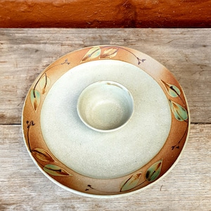 Può includere: Un piatto da portata in ceramica con una ciotola più piccola al centro. Il piatto ha uno sfondo beige chiaro con un motivo a foglie marroni e verdi.