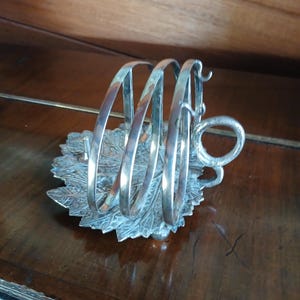 May include: A silver napkin holder with a leaf-shaped base and curved rings. The holder has a decorative handle and is made of metal. The design is vintage and elegant. The napkin holder is likely made of silver-plated metal.