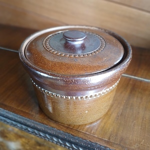 Puede incluir: Una olla de cerámica marrón con tapa. La olla tiene un diseño circular en la tapa y una forma cilíndrica con una banda decorativa de pequeños puntos en relieve. La olla está sobre una superficie de madera.