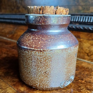 May include: A small, brown stoneware jar with a cork stopper. The jar has a textured surface and a rounded shape, with a darker brown band near the top. The cork stopper is a light brown colour and fits snugly in the jar's opening. The jar has a small, embossed design on the side.