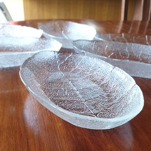 Puede incluir: Cuatro platos de servir de vidrio transparente con forma de hoja sobre una mesa de madera. Los platos son de forma ovalada y tienen una superficie texturizada que se asemeja a una hoja.