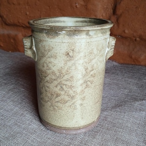 May include: A cylindrical ceramic vase with a speckled beige finish and two small, decorative handles. The vase has a slightly flared rim and a light brown base. The overall design is rustic and handmade, suitable for home decor.