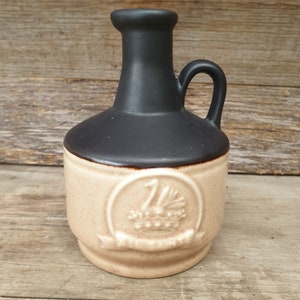 May include: A small ceramic jug with a black top and a beige bottom. The jug has a handle and a raised design of a swan with a crown on the front. The design is surrounded by a circle and a banner.