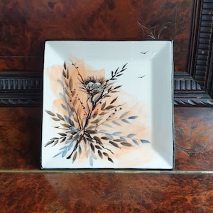 May include: A white square ceramic plate with a black rim, featuring a hand-painted design of a bird's nest with two eggs, surrounded by brown and blue leaves on a watercolor background.