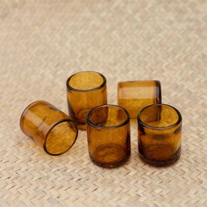 Puede incluir: Cinco vasos cilíndricos de color ámbar. Los vasos son de vidrio transparente y tienen un diseño simple y minimalista. Están dispuestos sobre una superficie tejida de color marrón claro. Los vasos están vacíos y listos para ser llenados.