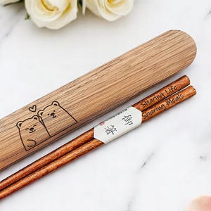 May include: Wooden chopsticks with a matching case. The case features a pair of bear illustrations with a heart above their heads. The chopsticks are brown with the words "Sharing Life Sharing Meals" printed on them. The chopsticks are in a white paper sleeve with Japanese text.