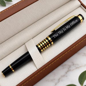 May include: A black and gold ballpoint pen with a gold clip and a decorative gold-banded grip section. The pen is engraved with the text "The Sky Is My Office" and is presented inside a brown rectangular case with a cream-colored interior.