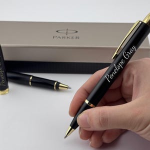 May include: A black ballpoint pen with gold-colored trim and a clip, featuring the name Penelope Gray engraved in white script. A matching pen cap and a Parker branded gift box are visible in the background, completing the set of writing instruments.