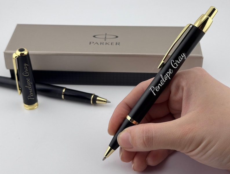 May include: A black ballpoint pen with gold-colored trim and a clip, featuring the name Penelope Gray engraved in white script. A matching pen cap and a Parker branded gift box are visible in the background, completing the set of writing instruments.