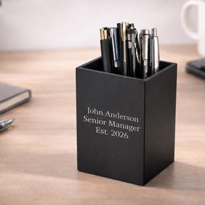 May include: A matte black square pen holder containing various pens. The front of the holder is engraved with the white text: John Anderson, Senior Manager, Est. 2026. The container is a functional desk accessory designed to hold writing instruments.