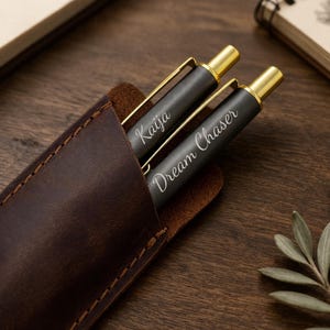 May include: Two black pens with gold accents and the inscriptions "Kaija" and "Dream Chaser" are displayed in a brown leather pen holder. The pen holder is on a wooden surface, with a notebook and pencil in the background.