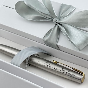 May include: A silver pen with gold accents and the engraved name "Chasyn Roberts" rests inside a white gift box. The box is tied with a silver satin ribbon, creating an elegant presentation.