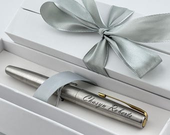 May include: A silver pen with gold accents and the engraved name "Chasyn Roberts" rests inside a white gift box. The box is tied with a silver satin ribbon, creating an elegant presentation.