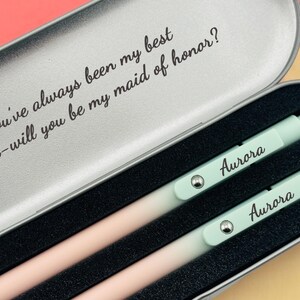 May include: A silver metal box with a message engraved on the lid: "You've always been my best friend - will you be my maid of honor?" The box contains two pastel green pens with the name "Aurora" engraved on them.