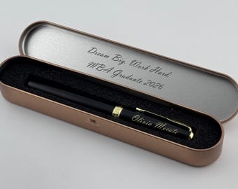 May include: A black pen with gold accents rests in an open, rose gold pen case. The pen case has a black interior and the lid is engraved with "Dream Big. Work Hard. MBA Graduate 2026." The pen is engraved with "Olivia Mecate."