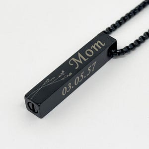May include: A black metal bar pendant necklace featuring an engraved floral design, the word Mom, and the date 03.05.57. The pendant is attached to a matching black metal chain with a box link style, and a small circular screw cap is visible at the base.