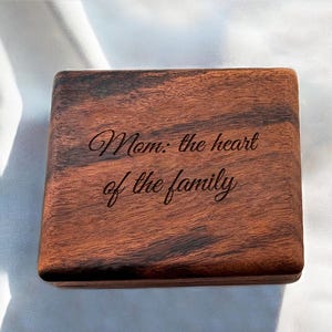 May include: A rectangular wooden box with a rich, dark brown finish. The lid is engraved with the words "Mom: the heart of the family" in an elegant script. The box is set against a white fabric background.