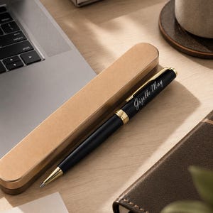 May include: A black ballpoint pen with gold-colored accents and a gold-colored clip, featuring the name Gizelle May engraved in white script on the barrel. The pen is positioned next to a matching gold-colored rectangular pen case.