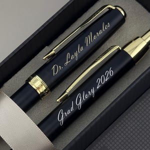 May include: A set of two matte black pens with gold-tone accents and clips, presented in a dark box. One pen is engraved with the text Dr. Layla Morales, and the other pen is engraved with the text Grad Glory 2026 in a cursive script font.