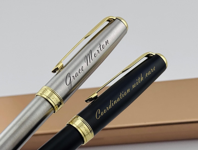 May include: Two elegant pens with gold accents. One pen is silver with the name "Grace Morton" engraved. The other pen is black with the phrase "Coordination with care" engraved. Both pens have gold clips and trim.