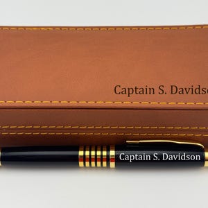 May include: A black ballpoint pen with gold-tone accents and the engraved text "Captain S. Davidson" rests in front of a rectangular tan leatherette case featuring yellow stitching and the same engraved name "Captain S. Davidson" on the lid.