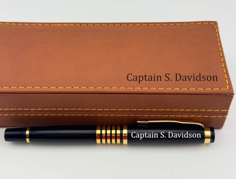 May include: A black ballpoint pen with gold-tone accents and the engraved text "Captain S. Davidson" rests in front of a rectangular tan leatherette case featuring yellow stitching and the same engraved name "Captain S. Davidson" on the lid.