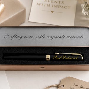 May include: A black pen with gold accents rests in a rose gold case lined with black foam. The case lid reads "Crafting memorable corporate moments." The pen is engraved with "Carl Radeveich."